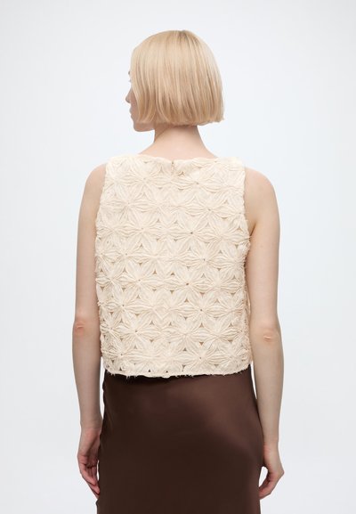 Femme aux cheveux blonds courts portant un haut beige texturé sans manches et une jupe marron foncé, vue de dos sur un fond uni.