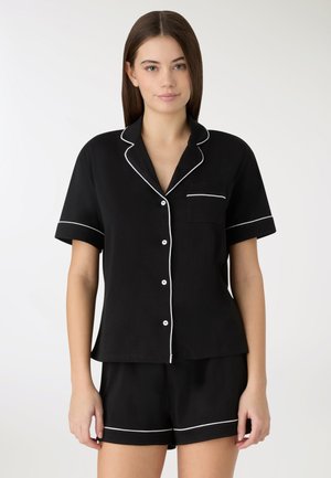 Młoda kobieta ubrana w czarną, krótka piżamę z guziczkami i dopasowane szorty z białą lamówką, stojąca na jednolitym tle.