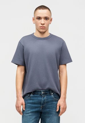 Νέος άνδρας με κοντά μαλλιά, φορώντας ένα απλό γκρι μπλουζάκι και μπλε τζιν, στέκεται μπροστά σε ένα ανοιχτό γκρι φόντο με ουδέτερο ύφος.