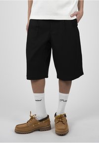 Personne portant un short noir au genou, des chaussettes blanches avec un texte noir et des chaussures à lacets beige, debout devant un fond uni.