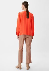 Orange Langarmbluse mit Puffärmeln, kombiniert mit braunen Hahnentritthosen und beigen, hohen Stiefeletten.