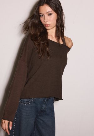 Jeune femme aux longs cheveux noirs portant un pull marron à épaules dénudées et un jean bleu avec des clous sur les côtés, debout devant un fond clair uni.