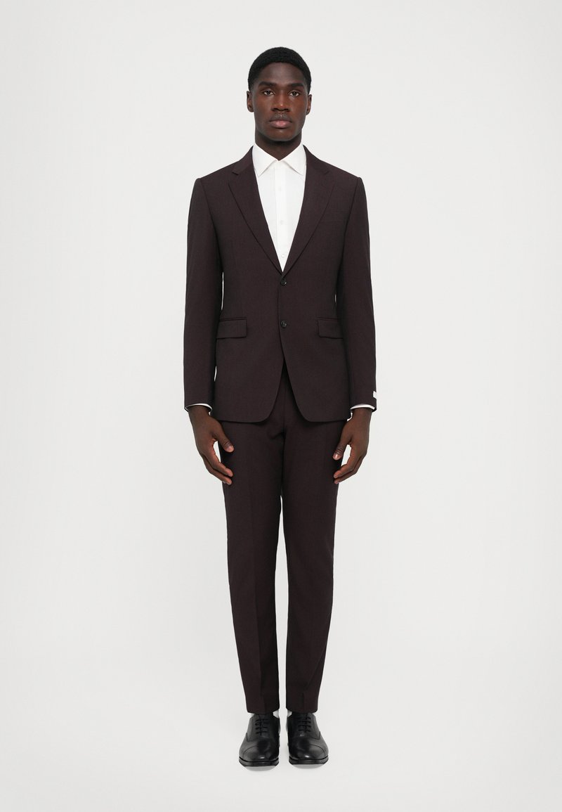 Suit bordeaux scuro con vestibilità su misura, rever a punta e due tasche frontali. Camicia bianca sotto, completato con scarpe in pelle nere.