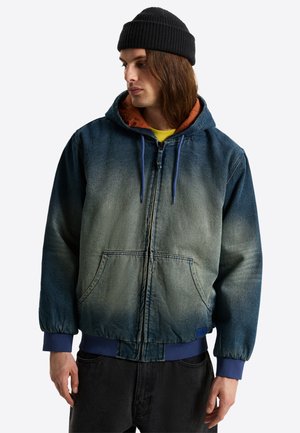 Ung mand iført en blå gradient denim lynlåshoodie med forlommer, en sort beanie og sorte bukser, kigger til venstre.