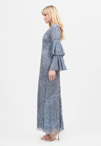 Długa, jasnoniebieska sukienka wieczorowa z wyszukanym haftem, dzwonkowymi rękawami, warstwowymi falbanami oraz płynącą spódnicą z falbaniastym dołem.