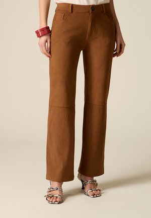 Brune bukser med høj talje og lige ben, båret med en hvid top, røde armbånd på venstre håndled og højhælede sandaler med åleskindsmønster.