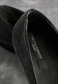 Black patent leather shoes with rounded toes, smooth texture, and stitched detailing. Inside shows a branded label and a star logo.