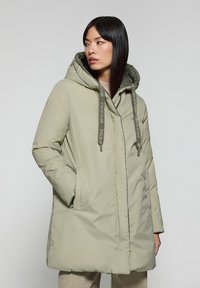 Ljusgrön puffjacka med huva, som har snoddar med stoppare, sidofickor och en mjuk, matt yta. Mellanlång design.