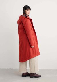 Parka lunga rossa con cappuccio, dotata di una chiusura a zip frontale, tasche laterali e un orlo leggermente curvo. Abbinata a pantaloni in velluto a coste color crema e stivali scuri.