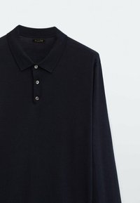 Chemise polo noire à manches longues avec trois boutons et un col rabattu, présentée sur un fond uni clair.