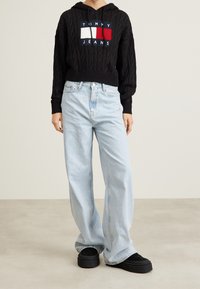 Person wearing black cable-knit Tommy Jeans hoodie, light blue wide-leg jeans, and black platform shoes standing on light floor.