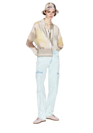 Młoda osoba nosząca beżowy kapelusz typu bucket z wzorem, przezroczystą pastelową koszulę, jasnoniebieskie spodnie cargo, beżową torbę na ramię i jasnoróżowe buty.