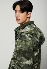 Zöld és szürke árnyalatú, álcázás mintás dzseki, kapucnival és patentos zárással. Könnyű, textúrált anyagból készült.