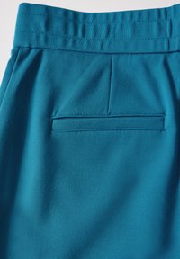 Teal rekbare stof met een gladde textuur, voorzien van een achterzak en een geplooide tailleband. Het ontwerp bevat subtiele plooien voor stijl.
