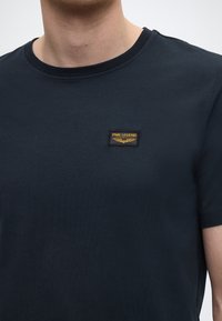T-shirt en coton bleu marine avec col rond et manches courtes. Présente un patch rectangulaire noir avec texte doré et emblème d'ailes.