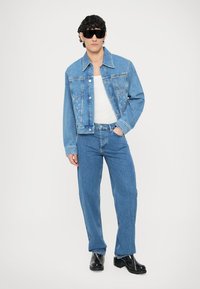 Jeans jakna svetlega pranja z vezenimi detajli, kombinirana s svetlo modrimi jeans hlačami. Oblečena z belo majico brez rokavov in črnimi škornji.