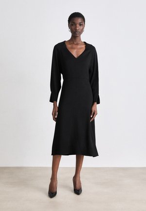 Black dress with a V-neck, long sleeves, and a flared hemline. Made of smooth fabric with a fitted waist. Worn with black heels.