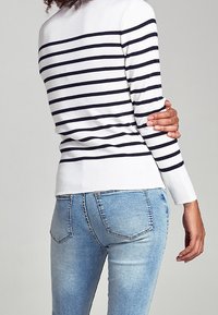 Persona che indossa una camicia bianca a maniche lunghe con strisce orizzontali nere e jeans di denim blu chiaro, in piedi su uno sfondo semplice.