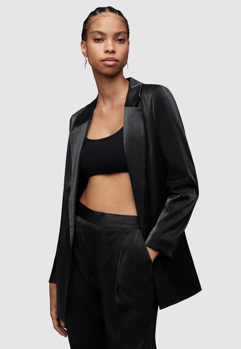 Schwarzer Satin-Blazer mit Reverskragen, kombiniert mit einem schwarzen Cropped-Top und passenden hochgeschnittenen Hosen, die glatte Texturen aufweisen.