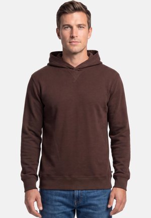Mann trägt ein schlichtes dunkelbraunes Kapuzensweatshirt und blaue Jeans, steht vor einem hellgrauen Hintergrund.