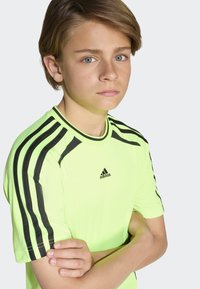 Neon gul Adidas t-skjorte med svarte skulderstriper og logo. Kortermet, tettsittende design, glatt tekstur og rund hals.