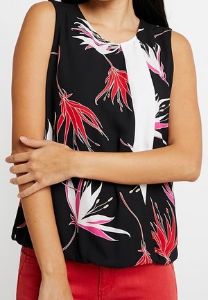 Czarny top bez rękawów z białym panelem na środku oraz czerwonymi, różowymi i białymi wzorami kwiatowymi, noszony z czerwonymi spodniami, ręce skrzyżowane na talii.