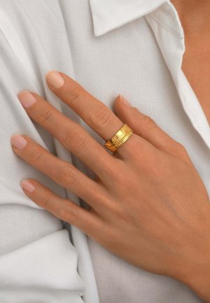 Main avec des ongles aux tons neutres portant une large bague en or texturée au majeur, reposant sur une chemise blanche à col.