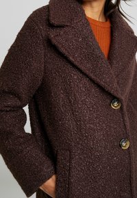 Brun texturerad kappa med notchkrage, dubbelknäppt design och stora knappar. Har sidofickor och ett fluffigt tyg.