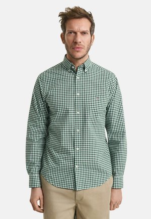 Mann steht nach vorne blickend, trägt ein grün-weiß kariertes Hemd mit Knöpfen und beige Hosen vor einem schlichten Hintergrund.