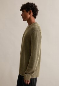 Olivově zelený žebrovaný svetr, dlouhé rukávy, kulatý výstřih, pletená textura, s volným střihem, nošený s tmavými kalhotami na neutrálním pozadí.