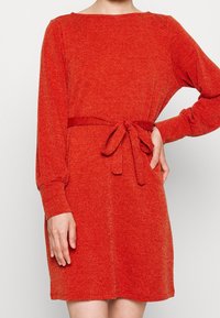 Vestido de punto rojo con mangas largas y un cinturón anudado en la cintura. Presenta un escote redondo y textura acanalada.
