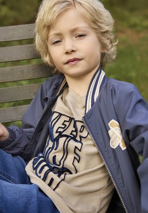 Blond dziecko siedzące na drewnianej ławce na zewnątrz, ubrane w niebieską kurtkę i beżowy sweter z dużymi ciemnymi napisami.