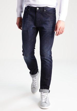 Uomo che indossa jeans blu scuro arrotolati e sneaker alte grigio chiaro, camminando in avanti su uno sfondo bianco.