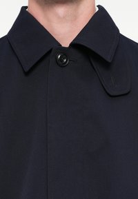 Tmavomodrý trenčkot so širokým golierom a jedným čiernym gombíkom na zapínanie. Vyrobený z hladkej, textúrovanej látky s precízne prešitými detailmi.