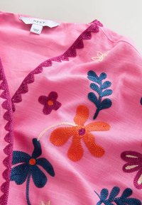 Pink fabric with embroidered blue, orange, and purple flowers and leaves, and dark pink scalloped trim along the neckline.