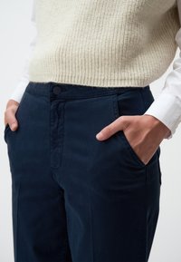 Marineblauwe, gep tailored broek met een rechte voorkant, zijzakken en een knoopsluiting. Het materiaal lijkt glad met een lichte glans.