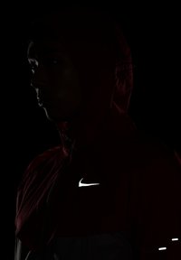 Veste à capuche Nike en tissu rouge et noir, avec un logo et des détails réfléchissants blancs. Le design met l'accent sur une silhouette moderne et élancée.