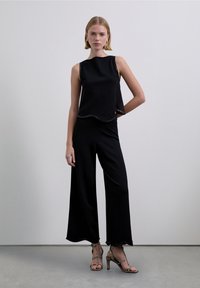 Top negro sin mangas con dobladillo festoneado, combinado con pantalones negros de pierna ancha. Tacones de tiras negros y dorados con acabado texturizado.