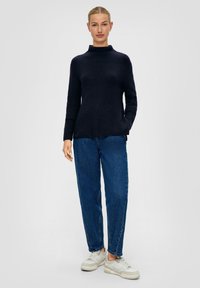 Marineblauwe gebreide trui met een hoge kraag, voorzien van lange mouwen en een ontspannen fit. Gecombineerd met hoge blauwe jeans en witte sneakers.