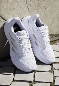 Paar weiße Adidas-Laufschuhe mit Mesh-Gewebe und strukturierten Sohlen, auf gepflastertem Steinboden vor einer strukturierten Wand platziert.