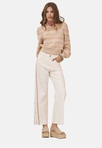 Suéter de punto beige claro con rayas rosas, combinado con pantalones de pierna ancha blancos que presentan botones laterales y un ribete decorativo. Sandalias de cuña beige.