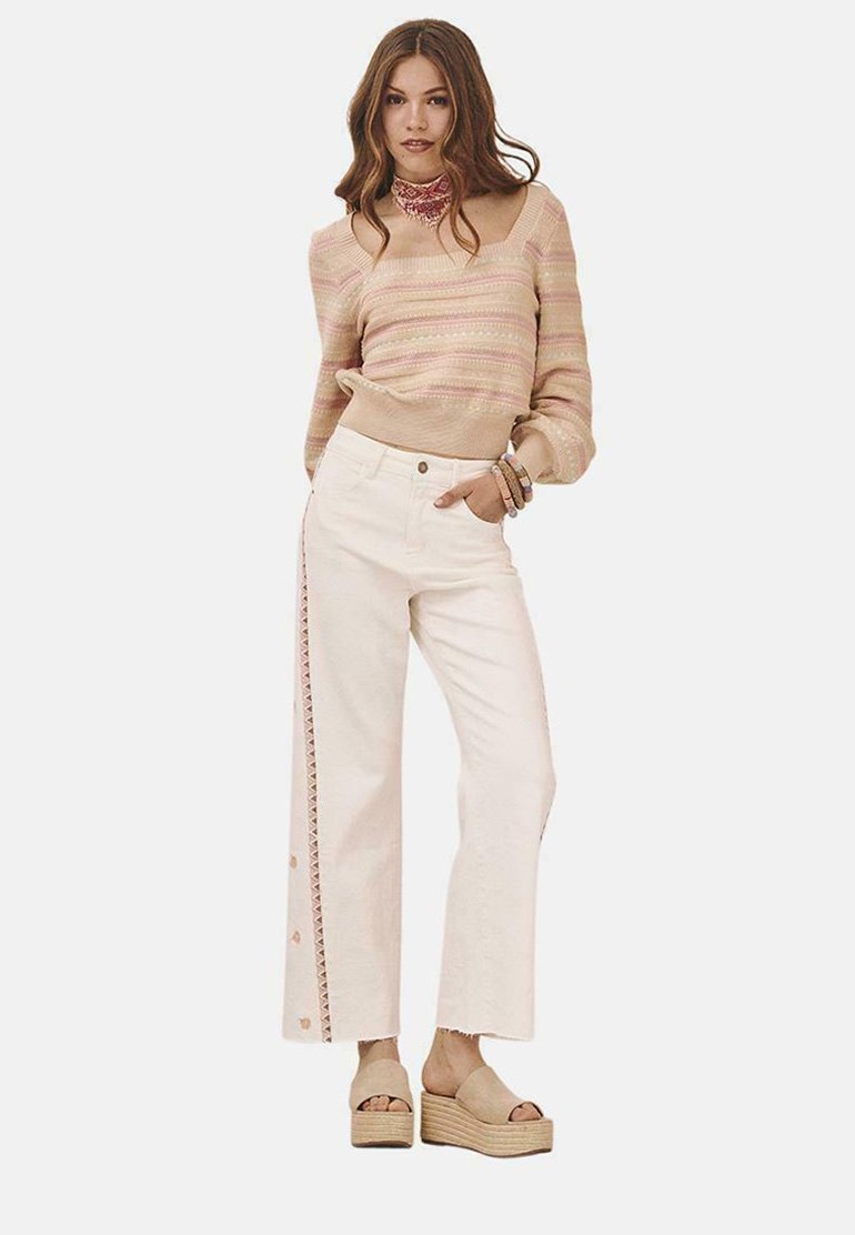 Suéter de punto beige claro con rayas rosas, combinado con pantalones de pierna ancha blancos que presentan botones laterales y un ribete decorativo. Sandalias de cuña beige.