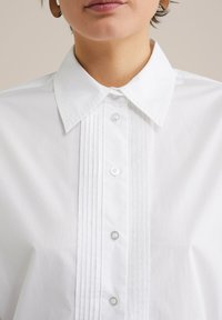 Biela košeľa s gombíkmi s klasickým golierom, plisovaným predným detailom a priehľadnými gombíkmi. Materiál vyzerá hladko a sviežo.