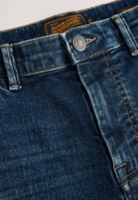 Nahaufnahme eines dunkelblauen Jeansbunds mit Metallknopf und braunem Etikett mit der Aufschrift Premium Denim Goods.