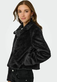 Jeune femme portant une veste en fausse fourrure noire avec col et un jean noir, posant avec une main dans la poche de la veste sur un fond uni.
