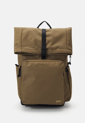 Brun kanvas ryggsekk med rull-toppslukking, justerbar svart stropp, frontlomme med glidelås, og sidelommer for organisering.