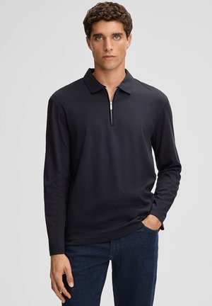 Mann in einem schwarzen Langarm-Polohemd mit Reißverschluss, das er mit einer dunklen Jeans kombiniert, steht mit einer Hand in der Tasche vor einem schlichten Hintergrund.