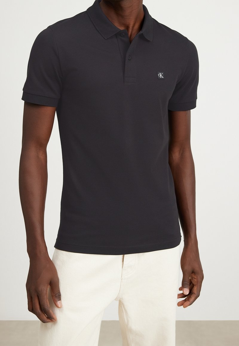 Homme portant un polo noir à manches courtes avec un petit logo sur la poitrine et un pantalon clair, debout devant un fond uni.