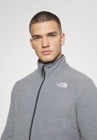 Hallhalli fleece jakk musta kaelusega, ees lukuga ja musta lukudetailiga. Sellel on väike valge logo vasakul rinnal.