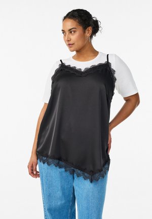 Žena nosi crni satenski kombinezon s ukrasnim čipkama preko bijele majice kratkih rukava i plavih traperica, stoji s jednom rukom na bok.